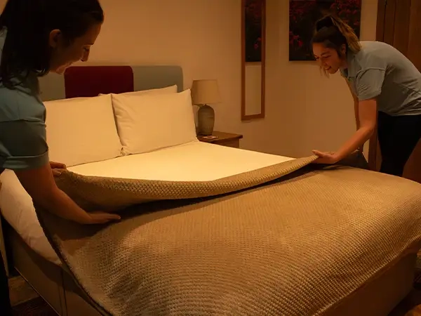Two women carefully arranging a bed throw on a meticulously made bed with crisp linen, creating a cozy and inviting atmosphere